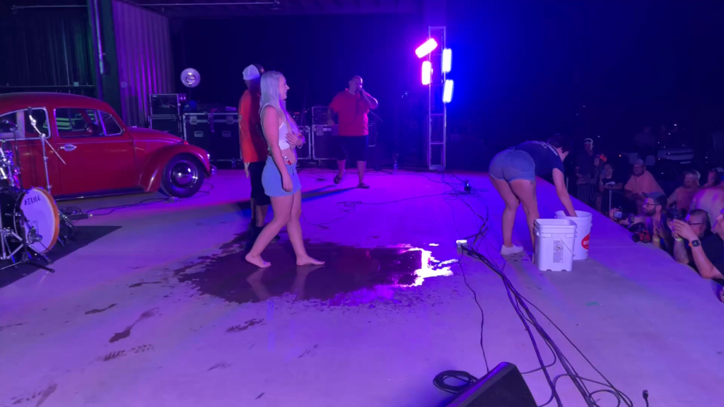 A woman in a blue tank top and shorts stands barefoot by a puddle, while another crouches with a bucket at a dimly lit stage.